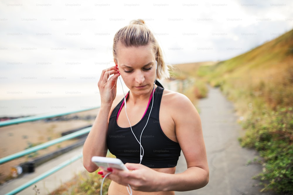 Jovem atleta corredora com fones de ouvido e smartphone em pé na praia na natureza, ouvindo música e descansando.