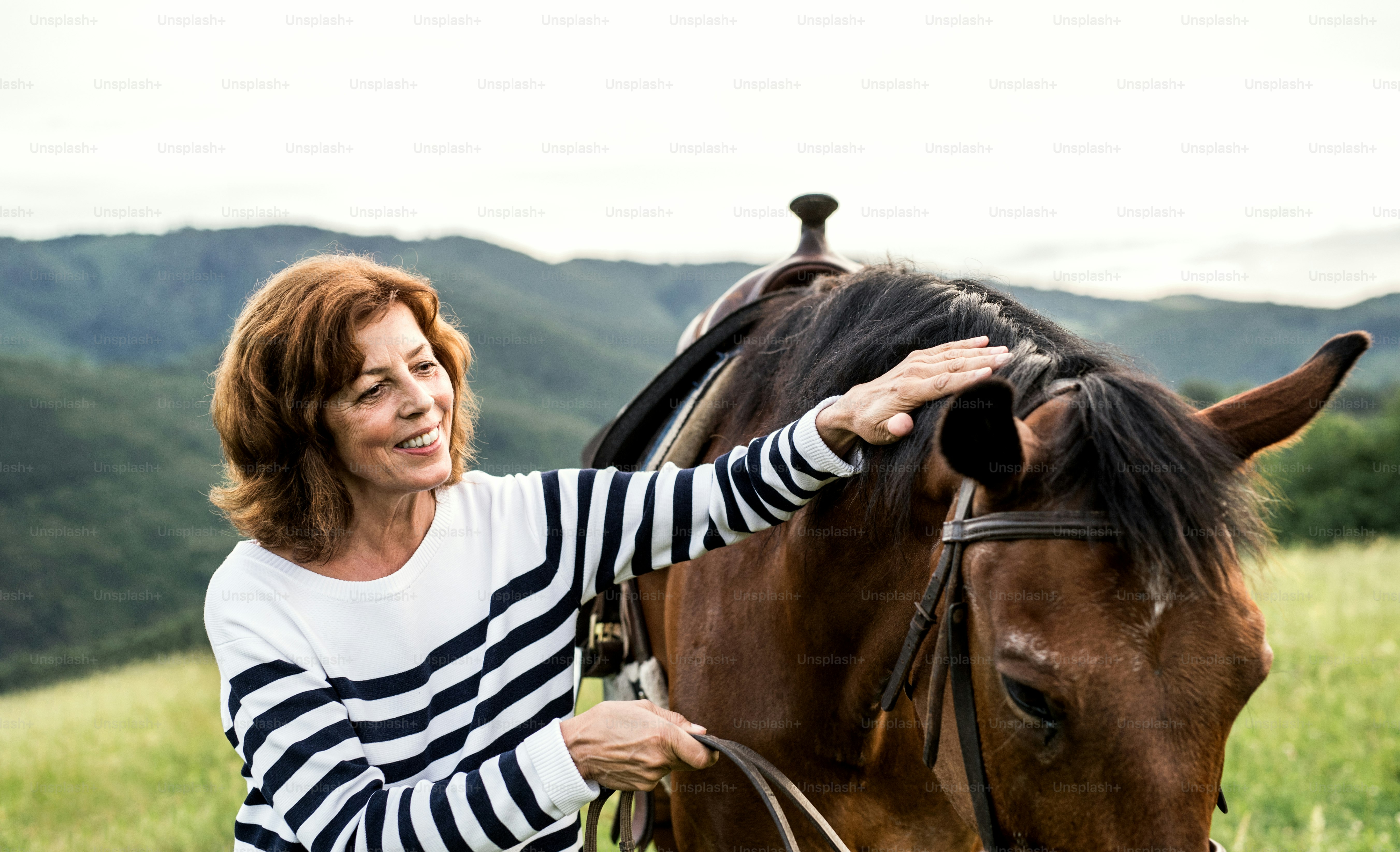Una mujer mayor feliz sosteniendo un caballo por su correa al aire libre en un pastizal.