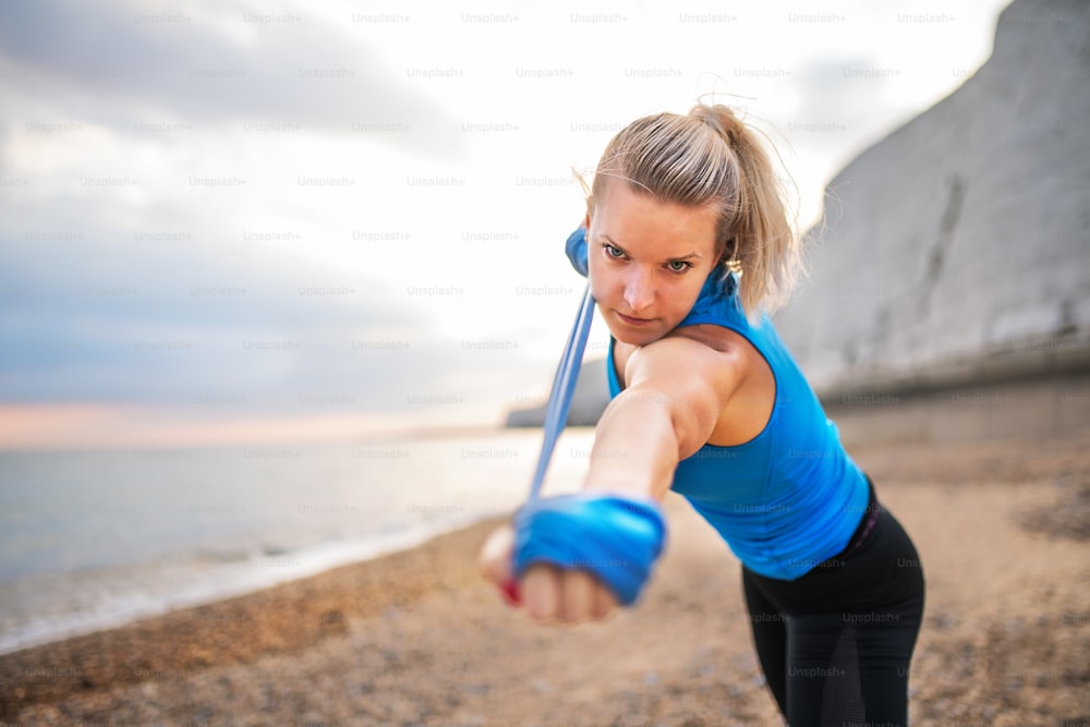 Young sporty woman runner doing exercise with elastic rubber bands outside on a beach in nature. Copy space.