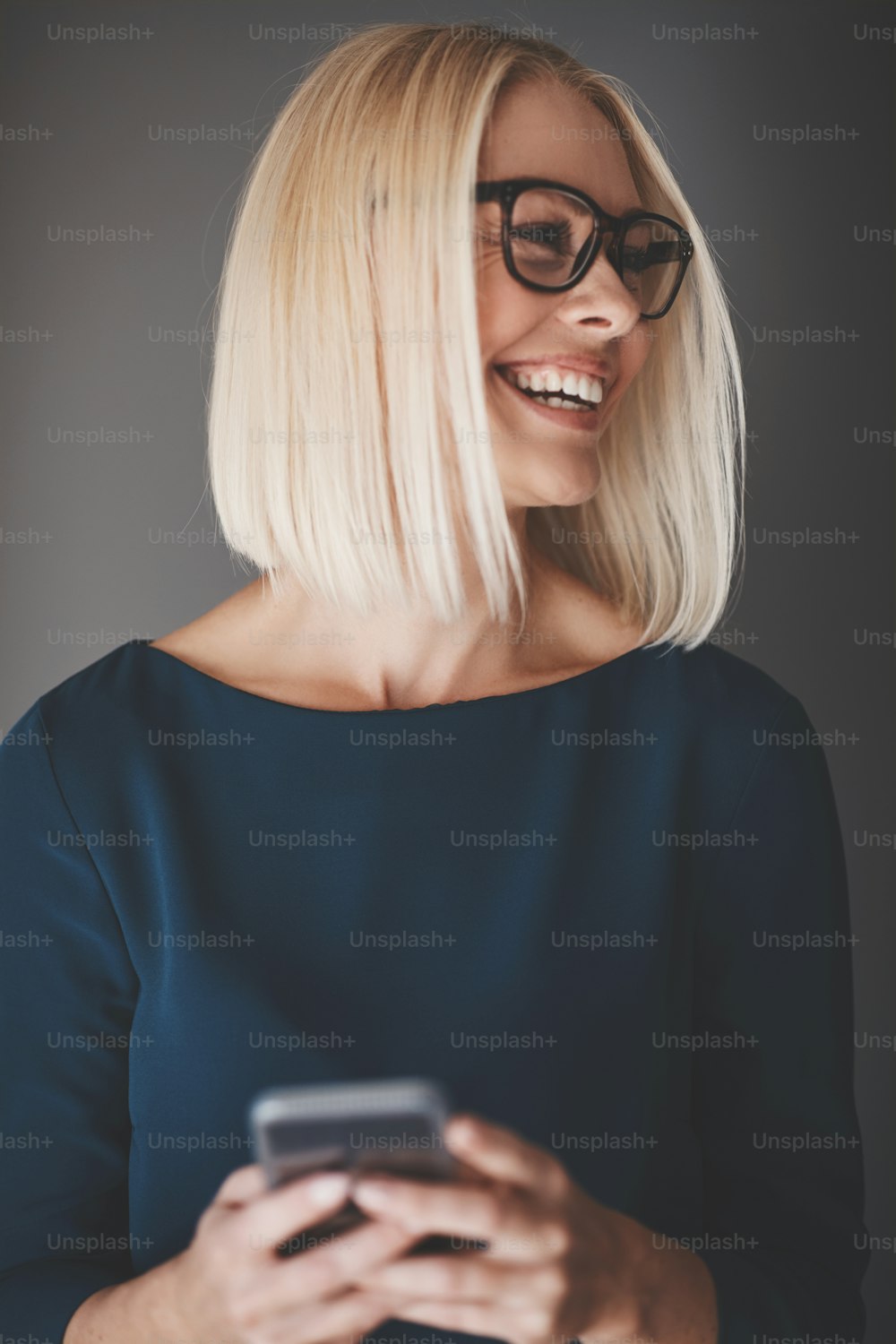 Young businesswoman reading a text message on her cellphone and laughing while standing against a gray background