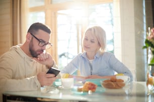 Hombre caucásico barbudo mostrando una imagen en el teléfono inteligente a su hermosa esposa mientras está sentado en la mesa de la cocina y desayuna