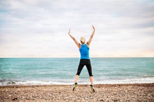 Giovane corridore donna sportiva in abbigliamento sportivo blu che salta fuori sulla spiaggia nella natura. Veduta posteriore.
