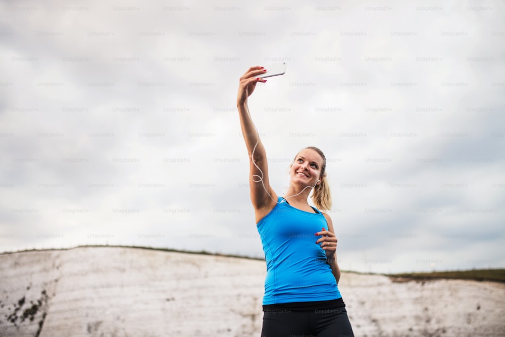 Jovem atleta mulher corredora com fones de ouvido e smartphone na praia, tirando selfie. Espaço de cópia.