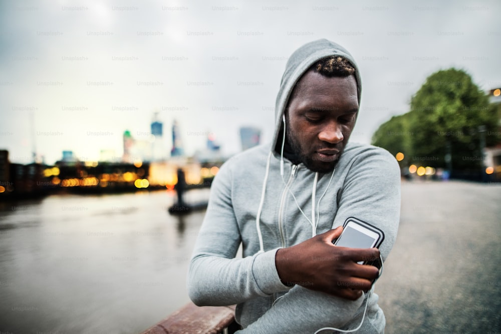 스마트워치, 이어폰, 스마트폰을 착용한 젊은 스포티한 흑인 남성이 도시의 다리에서 완장을 차고 휴식을 취하고 있다.