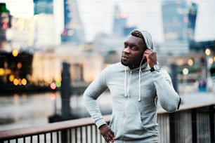 Young sporty black man runner with smartwatch and earphones on the bridge in a city, resting. Copy space.