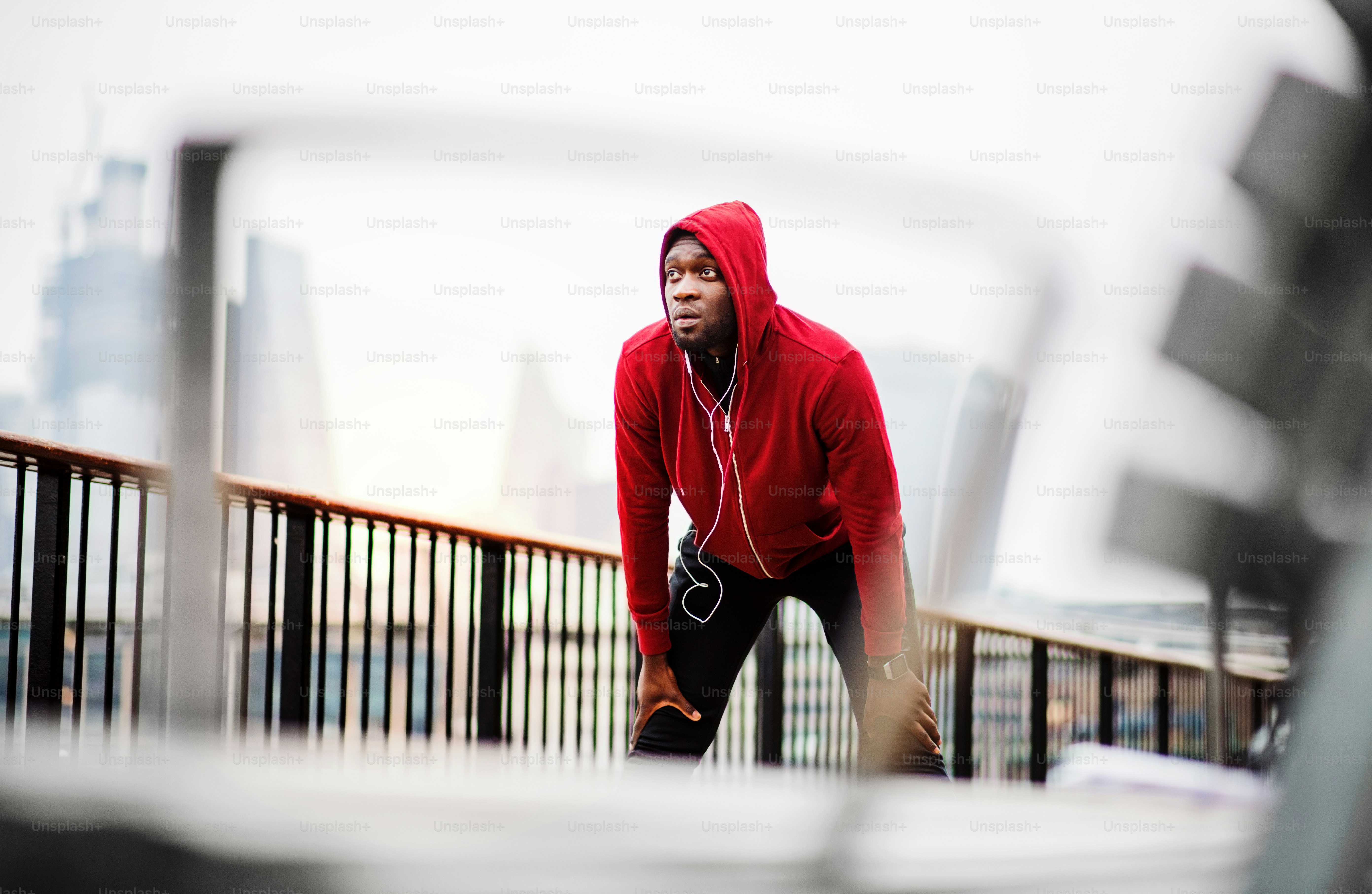A black man runner with earphones and red hood on his head standing on a bridge in a city, resting.