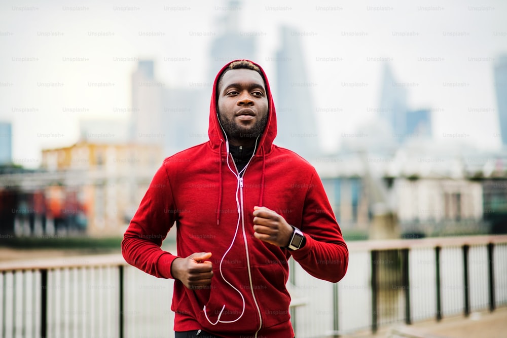 Jovem corredor negro esportivo com smartwatch e fones de ouvido correndo na ponte do lado de fora em uma cidade.
