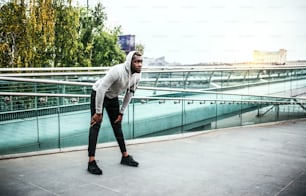 Young sporty black man runner with earphones and smartphone in an armband on the bridge in a city, resting. Copy space.