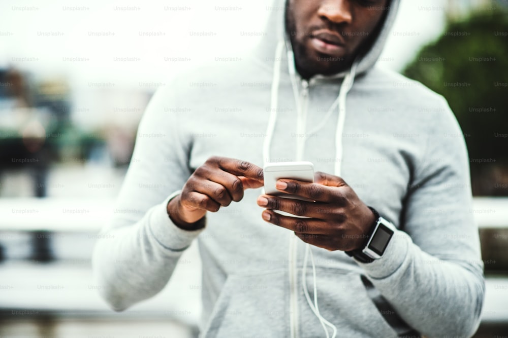Unkenntlicher junger sportlicher schwarzer Mann Läufer mit Smartwatch, Kopfhörern und Smartphone auf der Brücke in einer Stadt, ruhend. Speicherplatz kopieren.