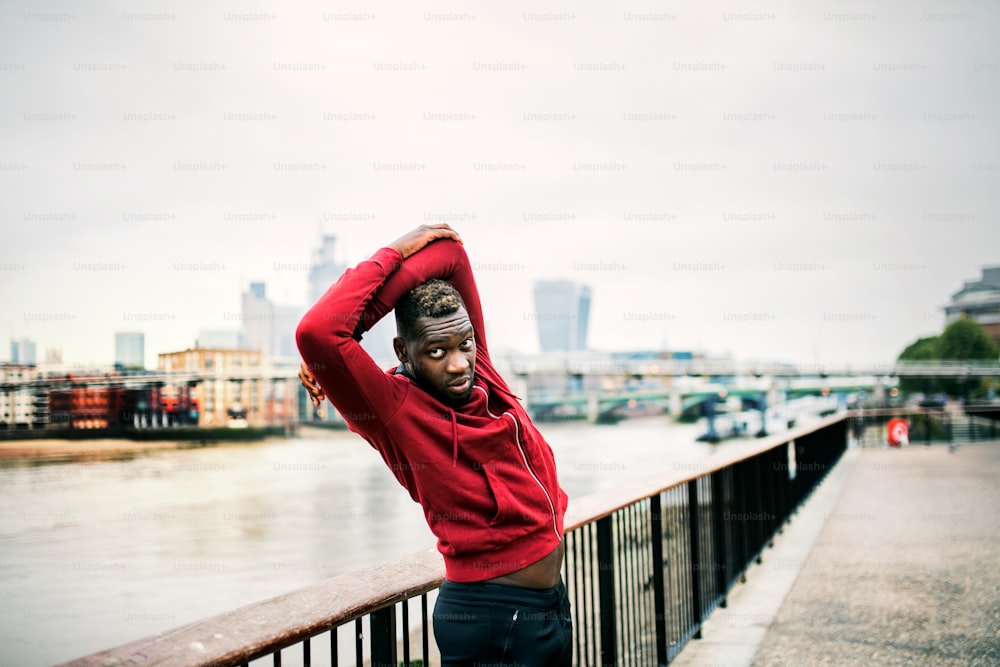 Young sporty black man runner with smartwatch doing stretching on the bridge outside in a city.