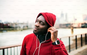 A close-up of black man runner with smart watch, earphones and hood on his head standing in a city. Copy space.