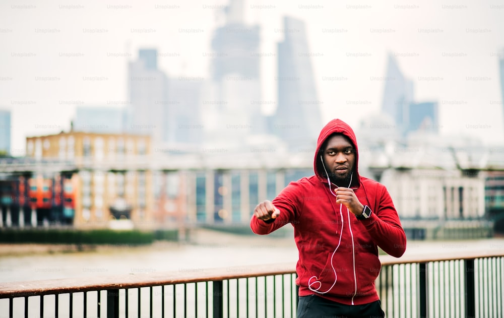 A black man runner with smart watch, earphones and hood on his head standing in a city in a boxing position. Copy space.