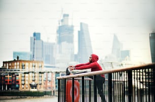 Jeune coureur noir sportif avec une montre connectée et des écouteurs étirant les jambes sur une balustrade sur le pont à l’extérieur dans une ville.