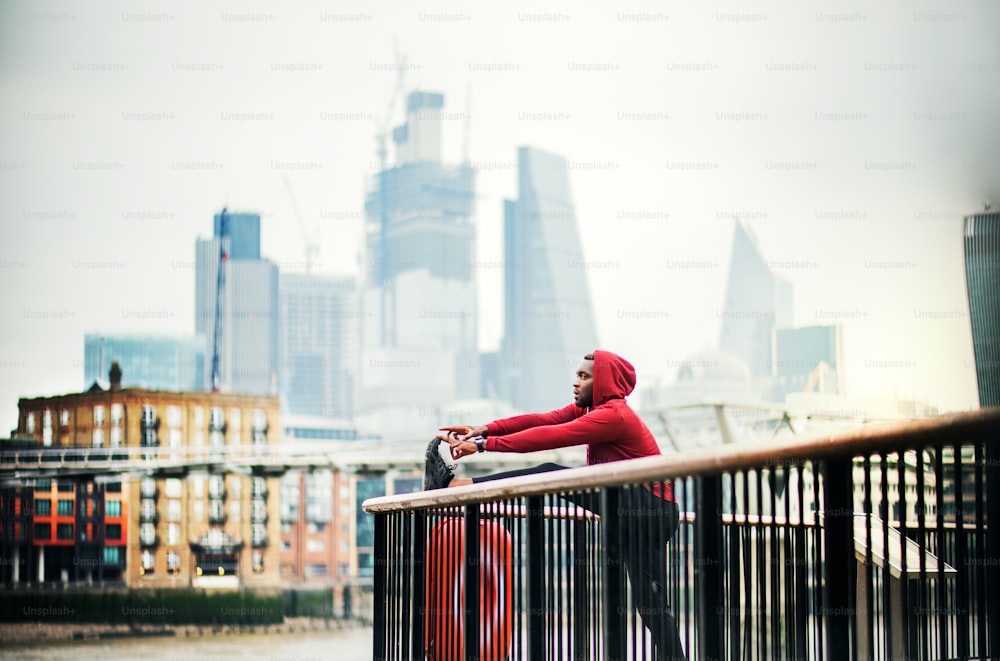 Junger sportlicher schwarzer Läufer mit Smartwatch und Kopfhörern, der die Beine auf einem Geländer auf der Brücke draußen in einer Stadt ausstreckt.