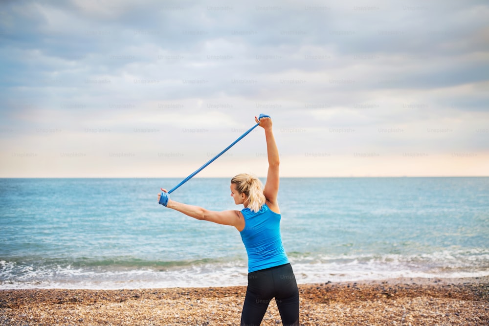 A rear view of young sporty woman runner doing exercise with elastic rubber bands outside on a beach in nature.