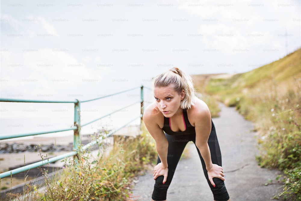 Young sporty woman runner in black activewear standing outside by the seaside, resting. Copy space.