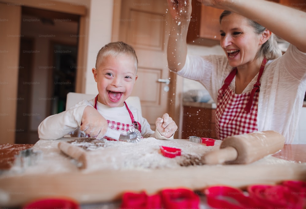 A laughing handicapped down syndrome child and his mother with checked aprons indoors baking in a kitchen, having fun.