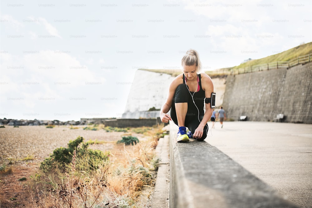 Giovane runner donna sportiva con auricolari e smartphone in una fascia al braccio fuori dalla spiaggia, allacciando i lacci delle scarpe.