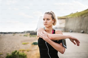 Giovane donna sportiva runner con auricolari e smartphone in fascia da braccio in piedi fuori sulla spiaggia nella natura, allungandosi.