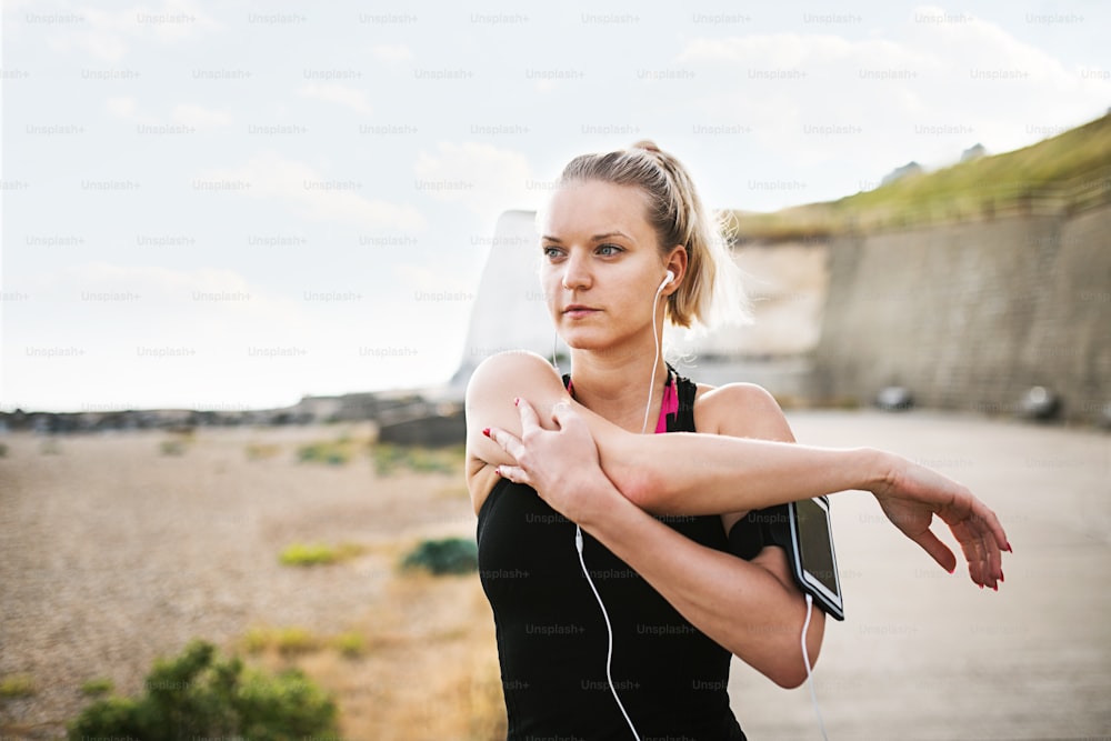 Young sporty woman runner with earphones and smartphone in armband standing outside on the beach in nature, stretching.