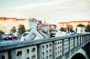 Ein fitter, reifer männlicher Läufer mit Kopfhörern, der sich draußen auf der Brücke in Prag streckt und Musik hört.