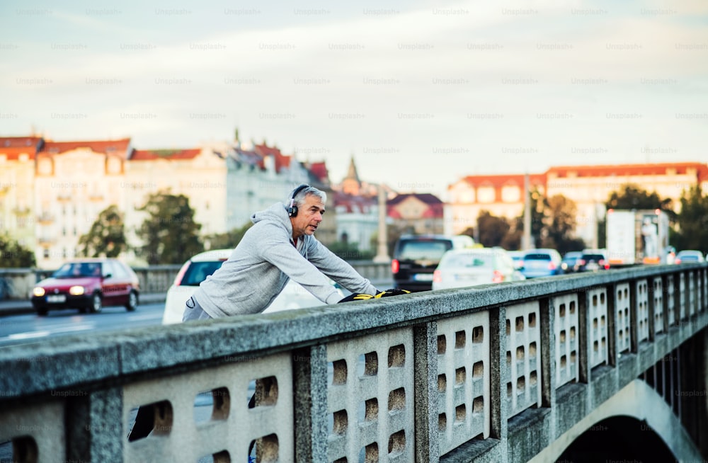 Un corredor masculino maduro en forma con auriculares estirándose al aire libre en el puente de la ciudad de Praga, escuchando música.