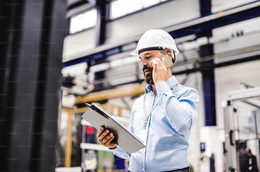 Ein Porträt eines reifen Industrieingenieurs mit Klemmbrett und Smartphone in einer Fabrik, der einen Anruf tätigt. Speicherplatz kopieren.