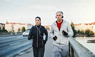 A fit sporty couple with headphones running outdoors on the bridge in Prague city, listening to music.