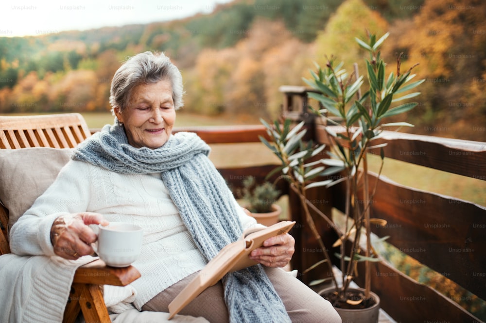 Una anciana mayor con una taza de café o té sentada al aire libre en una terraza en un día soleado de otoño, leyendo un libro.
