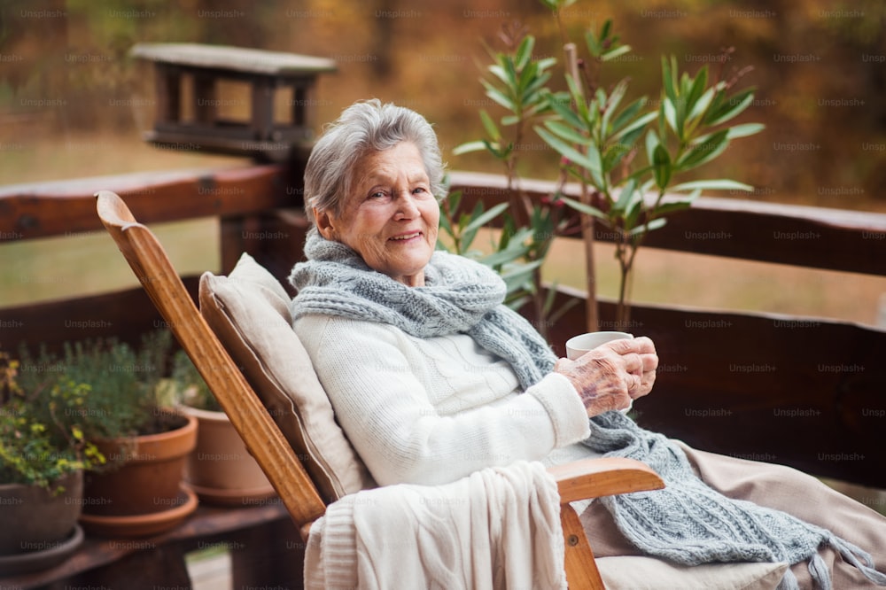 Une femme âgée avec une tasse de thé ou de café assise à l’extérieur sur une terrasse par une journée ensoleillée d’automne.