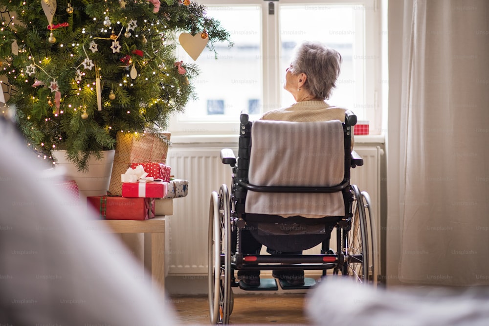 A rear view of a lonely senior woman in wheelchair at home at Christmas time, looking out of a window.