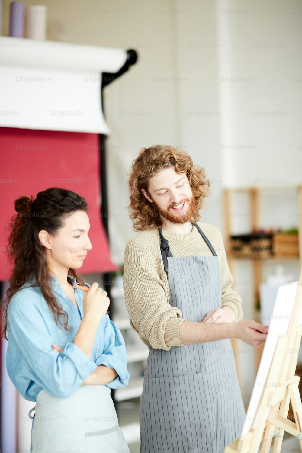 Young man in apron pointing at his groupmate painting on canvas while consulting at lesson