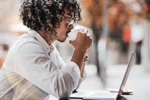 Um homem asiático barbudo pensativo com cabelos encaracolados e de óculos escuros está bebendo cuidadosamente seu delicioso chá enquanto está sentado na frente de seu laptop em um café ao ar livre durante uma pausa para o café