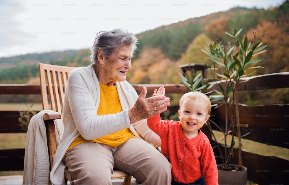 Eine ältere Frau mit einem Kleinkind Urenkel auf einer Terrasse an einem sonnigen Tag im Herbst, gibt High Five.