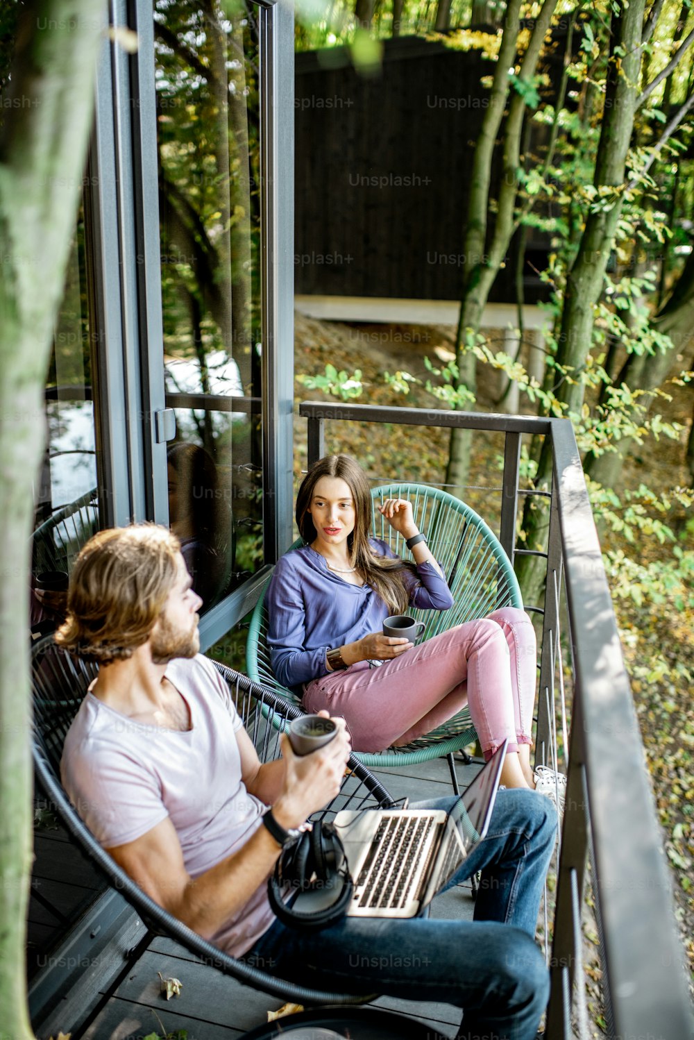 Hombre y mujer jóvenes que se relajan con el ordenador portátil y el café sentados en el balcón en el hermoso bosque verde