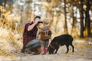 Un père mûr avec un chien noir et un fils en bas âge dans une forêt d’automne, utilisant des jumelles.