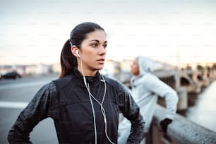 A fit young female runner with earphones standing outdoors on the bridge in Prague city, listening to music.