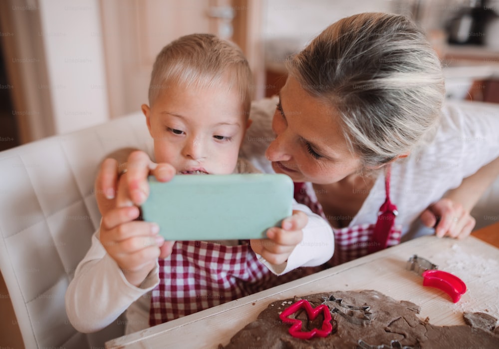 Ein behindertes Down-Syndrom-Kind und seine Mutter mit Smartphone drinnen in einer Küche, Selfie beim Backen.