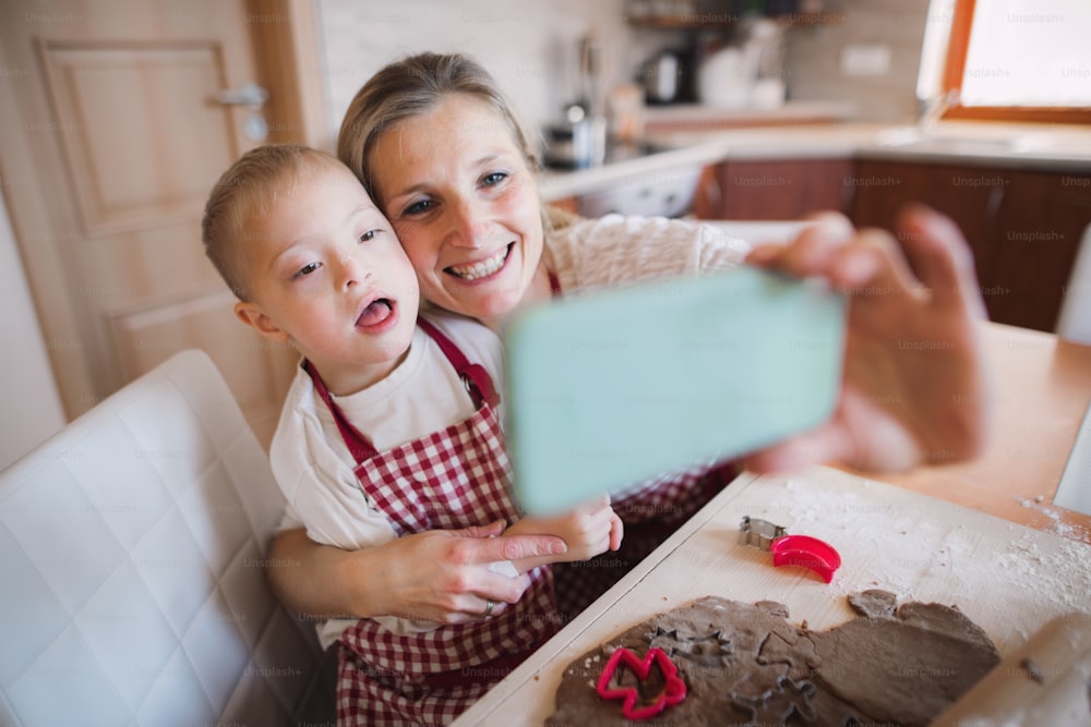 Un niño discapacitado con síndrome de down y su madre con un teléfono inteligente en el interior de una cocina, tomándose una selfie mientras hornean.