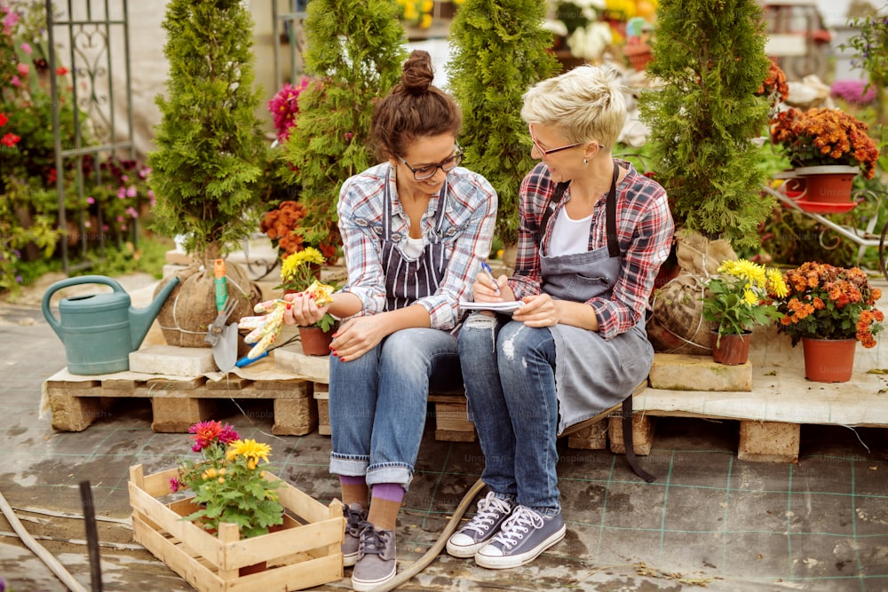 Two florist women sitting and writing in notebook their selling. All around potted flowers and plants.