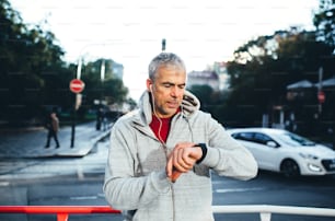 A portrait of an active mature man with earphones standing outdoors in city, checking the time.
