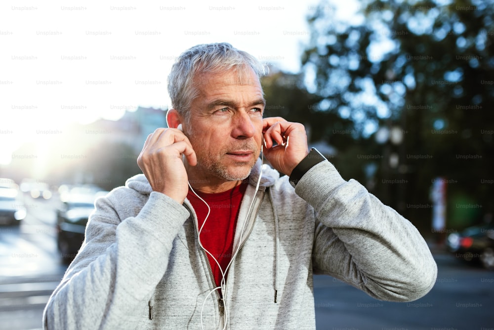 A portrait of an active mature man standing outdoors in city, putting earphones in his ears.