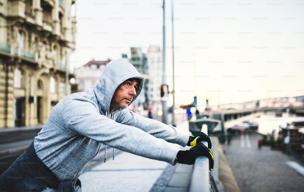 A fit mature male runner stretching outdoors on the street in Prague city, hood on his head.