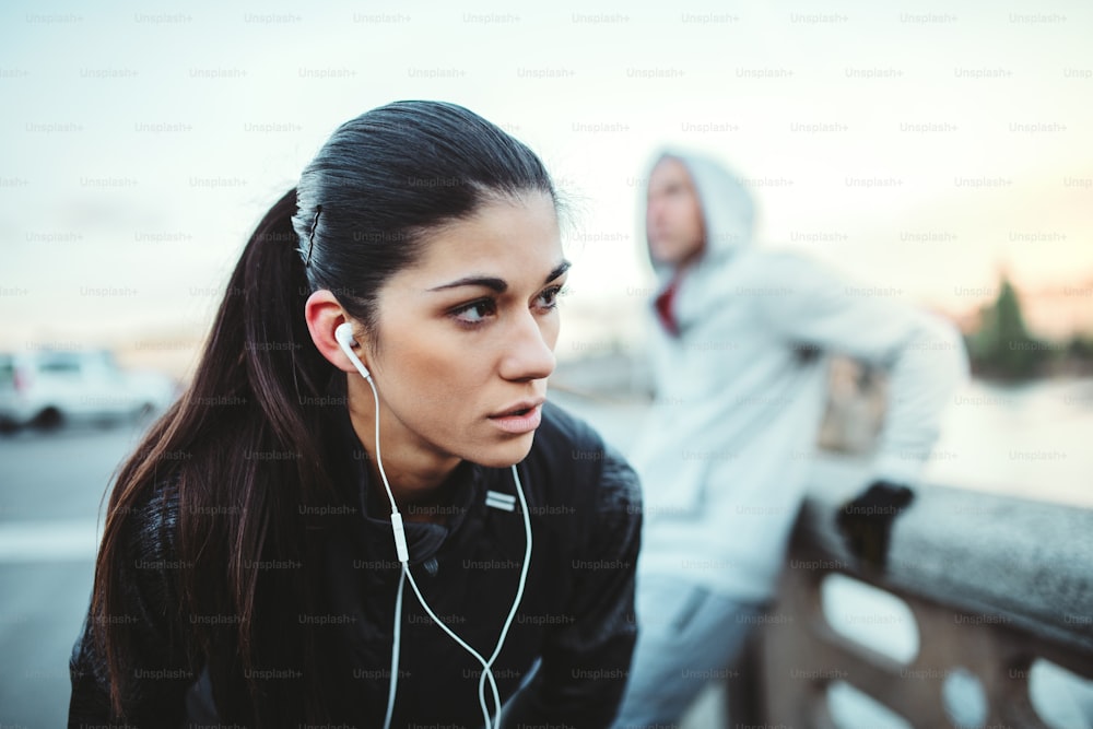 A fit young female runner with earphones standing outdoors on the bridge in Prague city, listening to music.