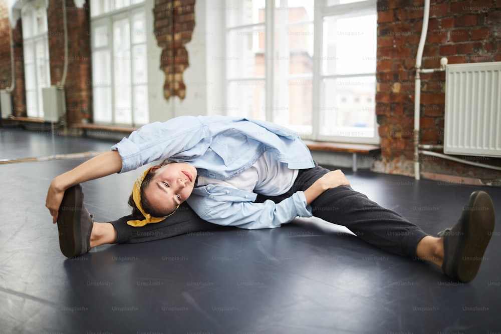 Young active female doing stretching exercise for arms and legs while sitting on the floor in studio