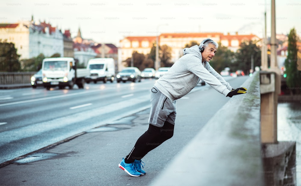 A fit mature male runner with headphones stretching outdoors on the bridge in Prague city, listening to music.