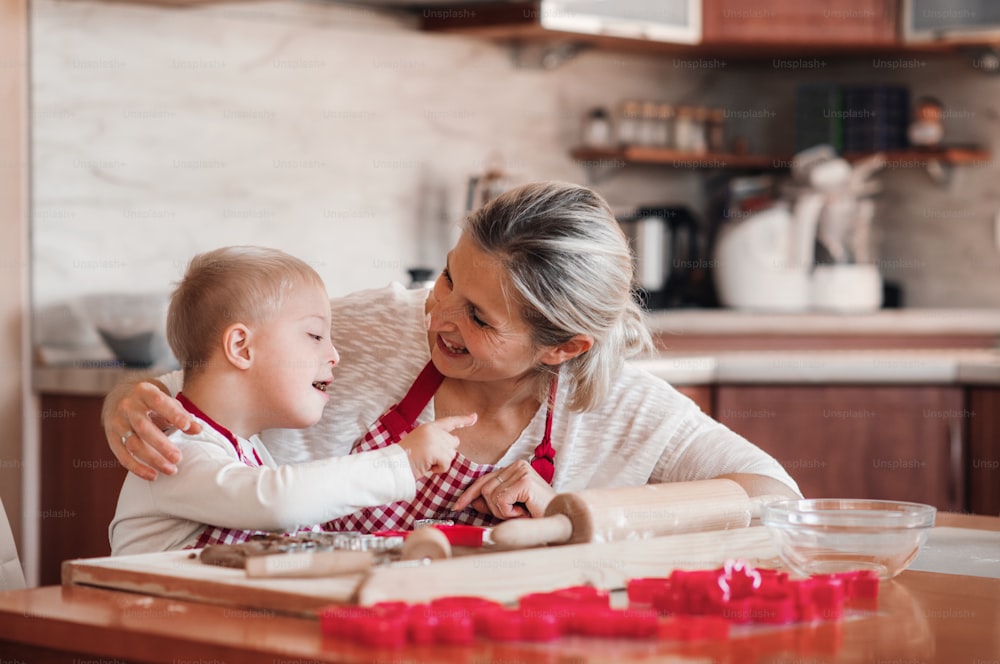 Un feliz niño discapacitado con síndrome de down y su madre con delantales a cuadros en el interior horneando en una cocina, divirtiéndose.