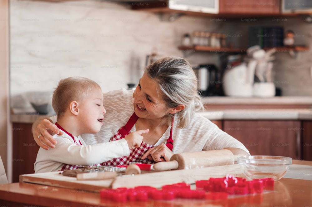 A happy handicapped down syndrome child and his mother with checked aprons indoors baking in a kitchen, having fun.