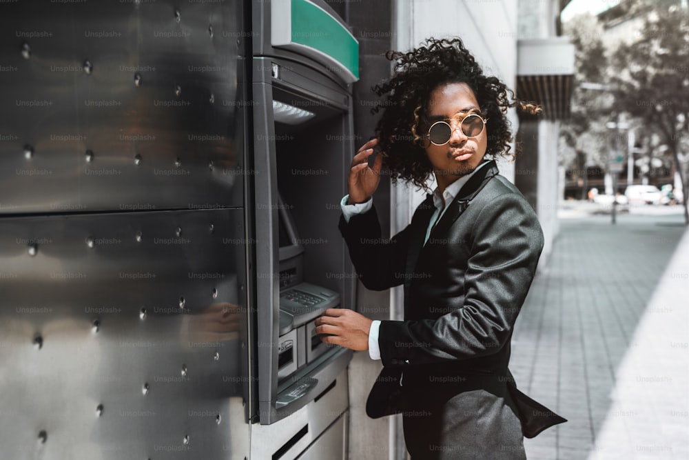 A curly Asian guy in fancy sunglasses and with curly hair is using an outdoor automated teller machine to replenish his deposit; man entrepreneur in business suit using ATM on the street