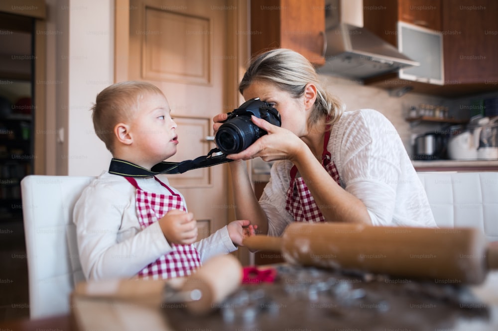 Un garçon handicapé trisomique et sa mère à l’intérieur dans une cuisine prennent des photos avec un appareil photo numérique tout en cuisinant.